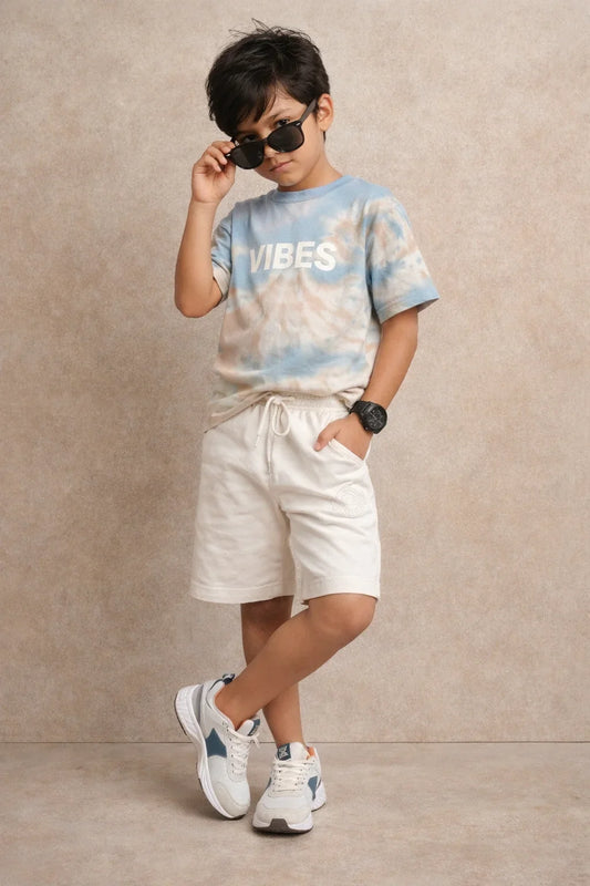 Stylish boy in tie-dye VIBES t-shirt, cream shorts, and sneakers posing with sunglasses