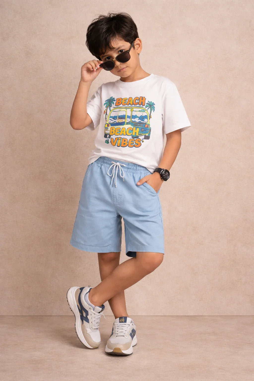 Stylish boy in beach vibes t-shirt, light blue shorts, sneakers, and sunglasses posing casually